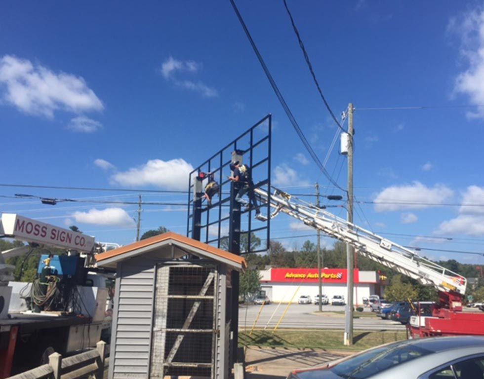 Workers on a lift installing a sign frame, a Moss Sign Co. truck is nearby, and a Johnson Auto Parts store is visible in the background under a blue sky.
