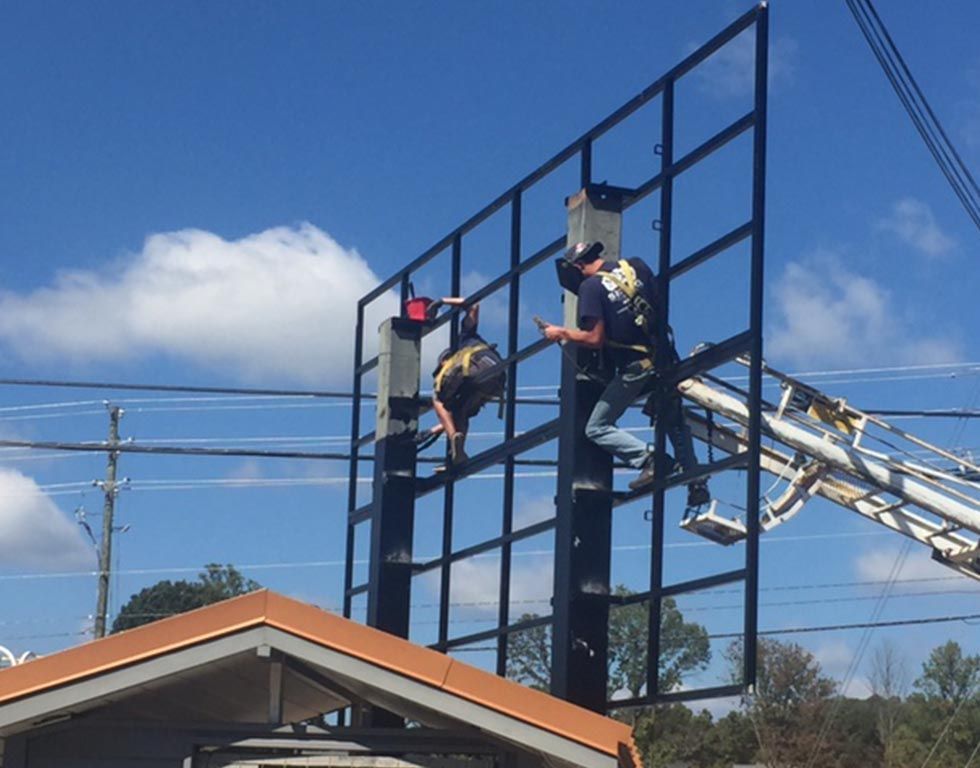 Two workers on a tall billboard frame, likely installing signage. One is suspended, the other is working from a lift, both wearing safety harnesses against a blue sky.