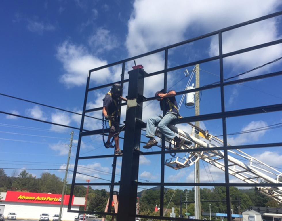 Two workers on a tall billboard frame wearing safety harnesses. The frame is black against a blue sky with clouds, near a Advance Auto Parts store.