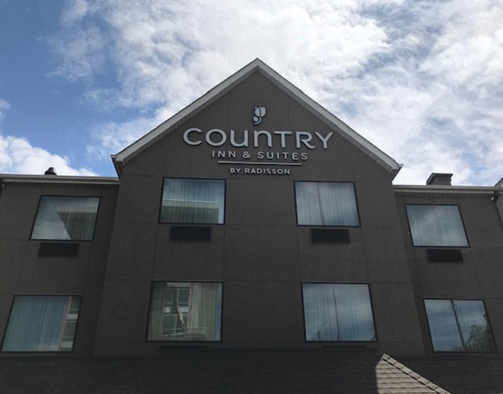 Country Inn & Suites by Radisson hotel exterior with a gray facade and the hotel's logo. Windows with curtains line the building under a bright blue sky.
