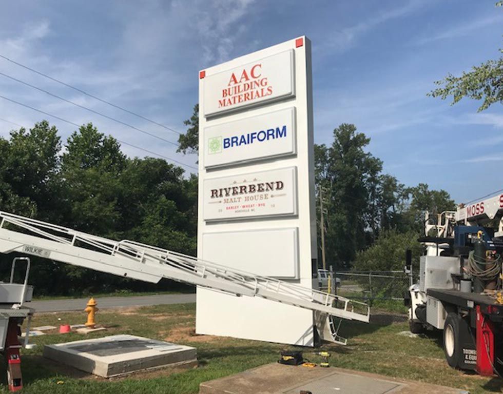 A tall, white signpost with four panels displaying company names: AAC Building Materials, Braiform, Riverbend. A lift truck is positioned next to the sign.