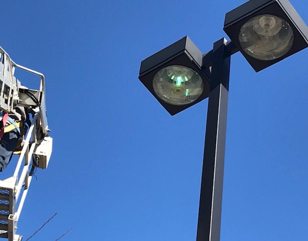 Ladder truck reaching up to a tall light pole with two lamps against a clear blue sky. A person is visible on the ladder.