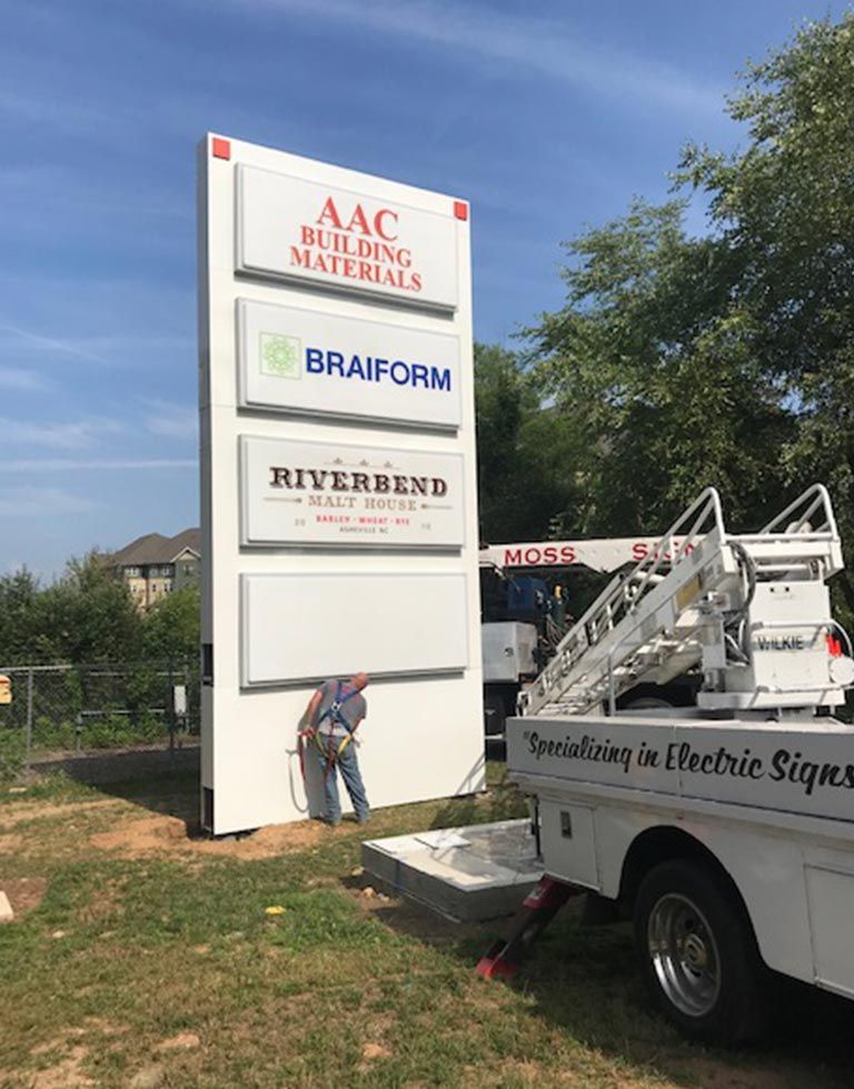 A worker adjusts a sign for multiple businesses mounted on a tall white pillar. The signs have the names AAC Building Materials, Braiform, and Riverbend.
