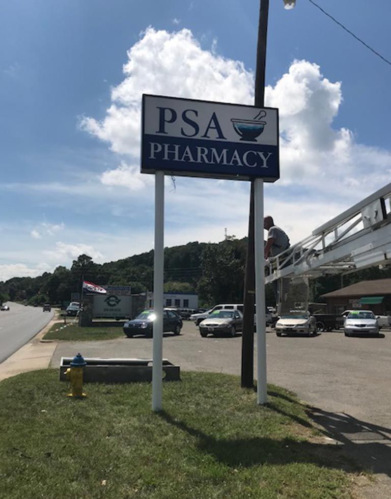 Sign for PSA Pharmacy with a person working on it, next to a road on a sunny day.