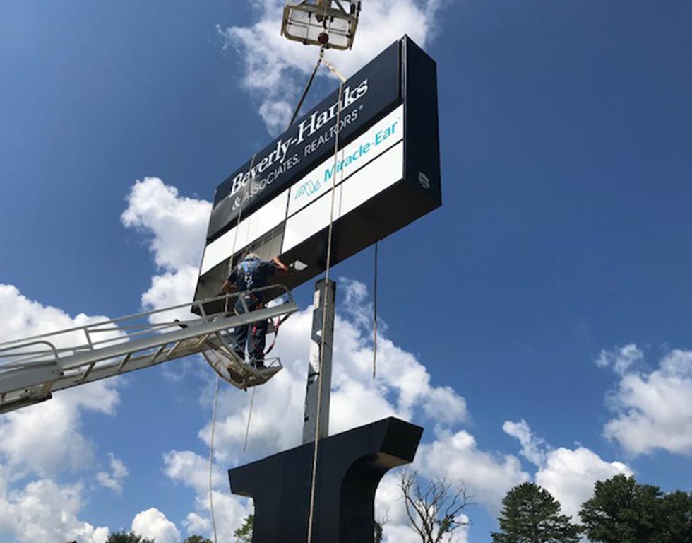 Workers on a lift install a sign for Beverly-Hanks Realtors and Miracle-Ear against a blue sky. The sign is black with white lettering.