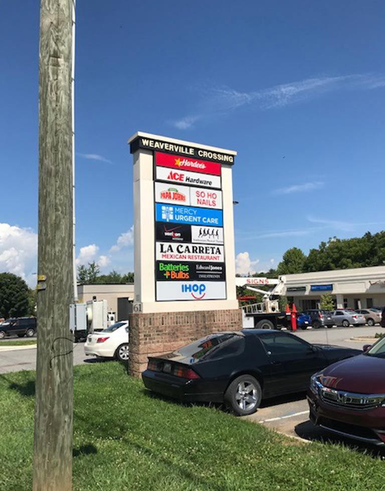 Signpost for a shopping center, with names of businesses listed, including a JCPenney, on a sunny day. A black car is parked in front.