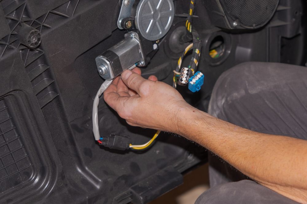 Person working on a car door, attaching a wire to a window motor. Black interior and visible wires.