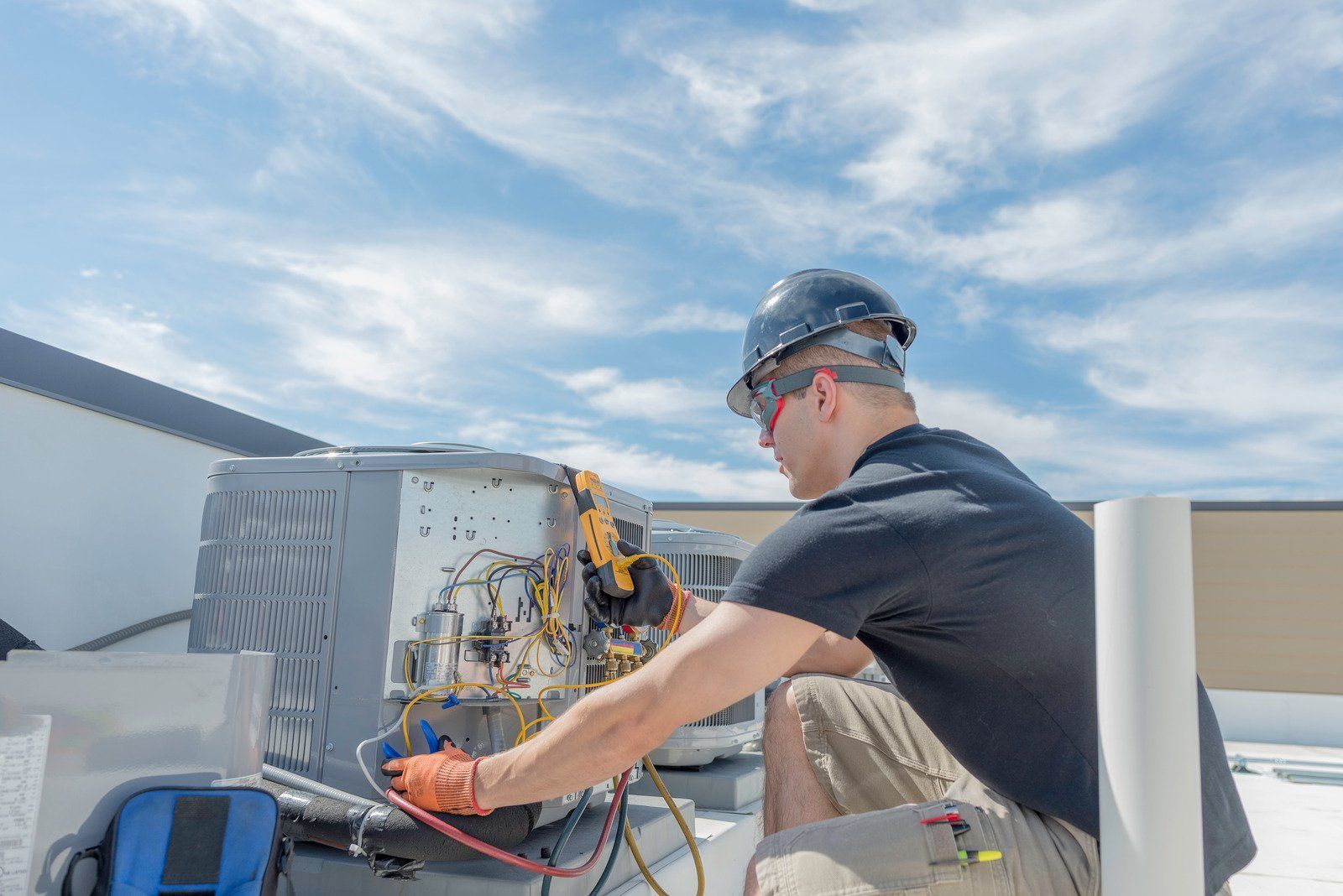 HVAC technician in a hard hat, repairing an air conditioning unit on a rooftop under a blue, cloudy sky.