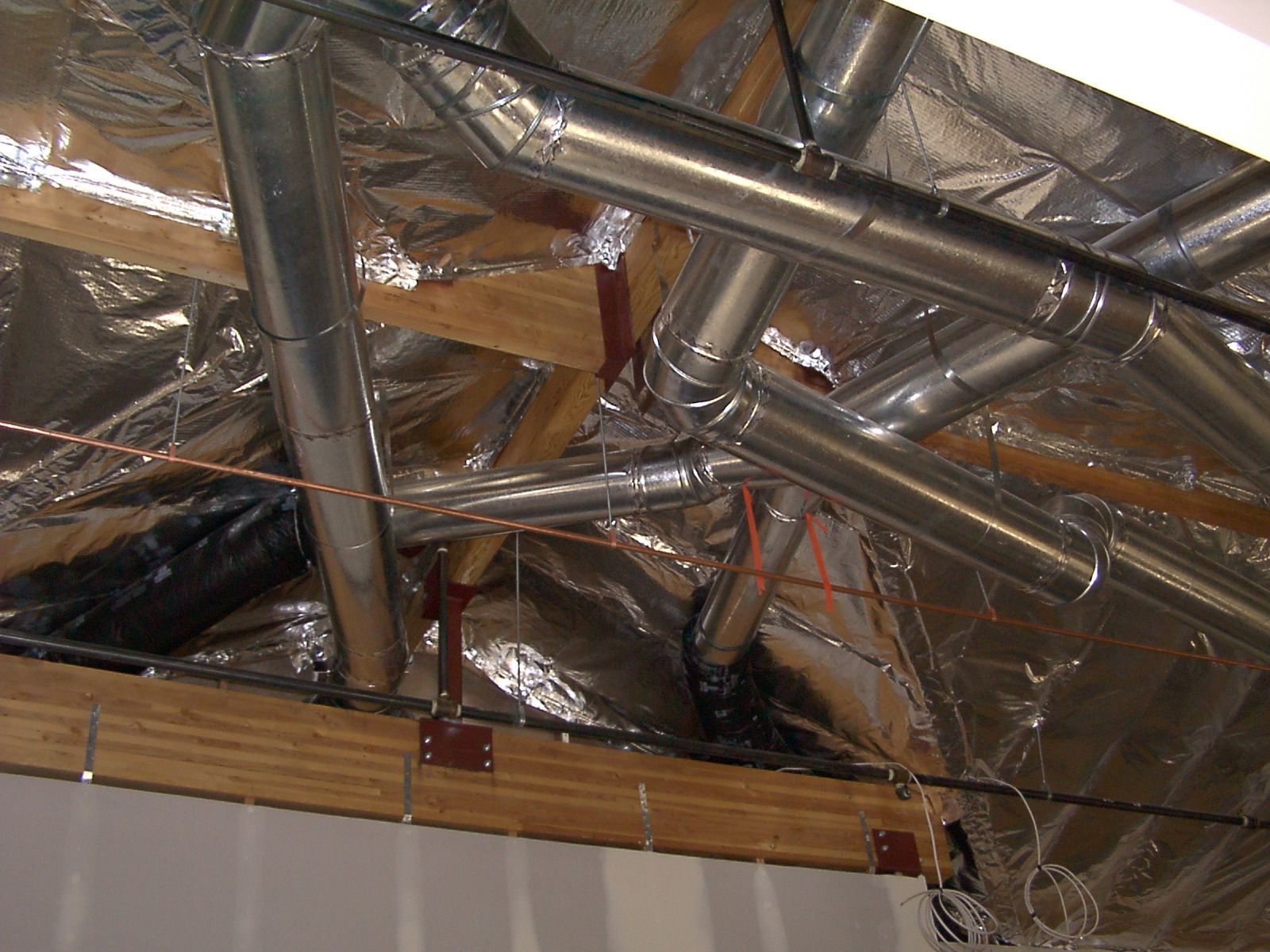 Silver HVAC ductwork installed within a wooden roof truss structure, insulated with reflective material.
