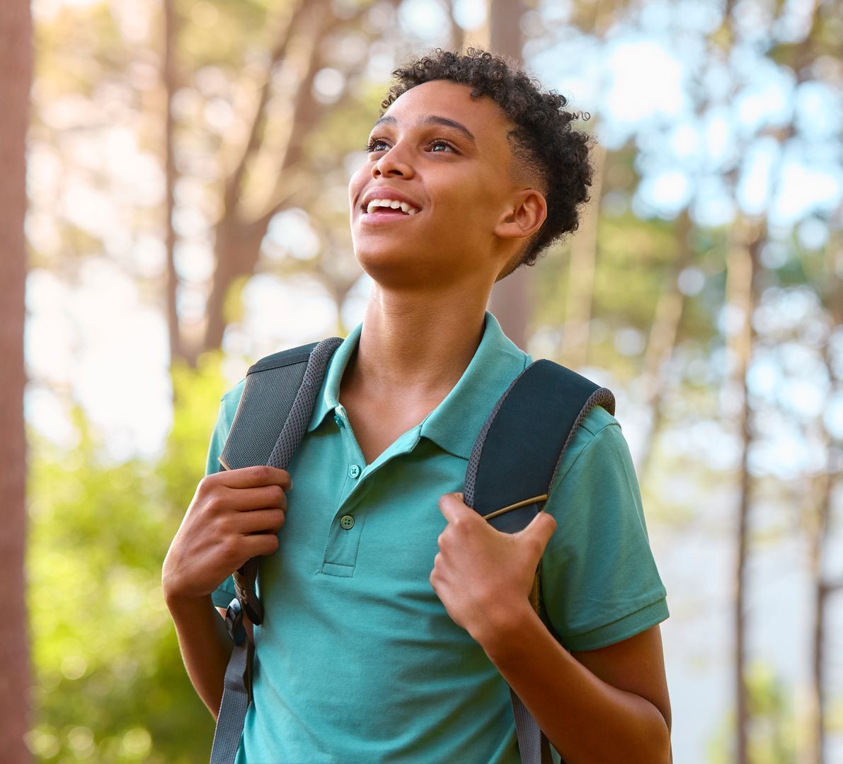 A young boy with a backpack is standing in the woods and smiling.