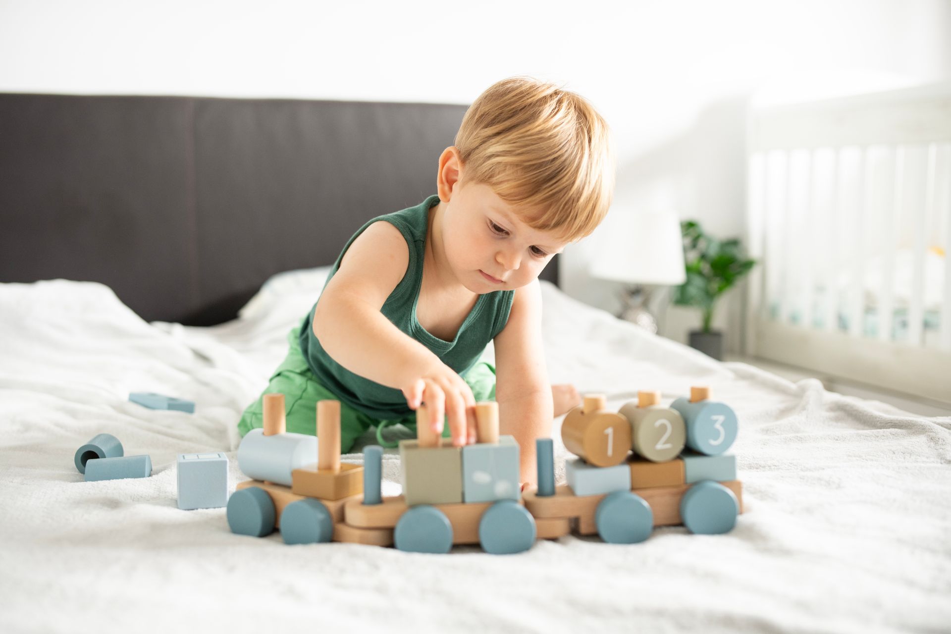 A little boy is playing with a wooden train on a bed.