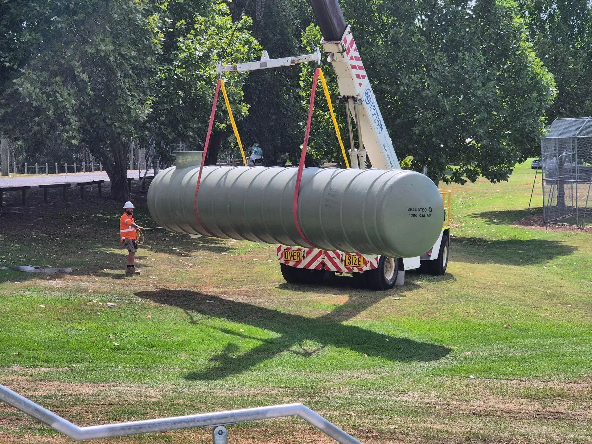 Large gray cylindrical tank being lifted by a crane in a grassy park, with workers nearby. Large gray cylindrical tank being lifted by a crane in a grassy park, with workers nearby.