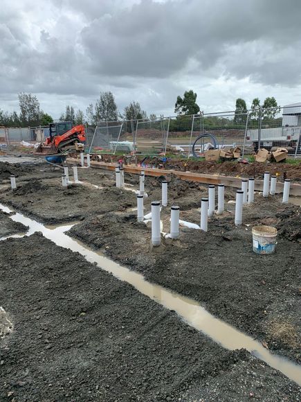 Construction site with gravel, white pipes, muddy trenches, and an excavator under cloudy skies