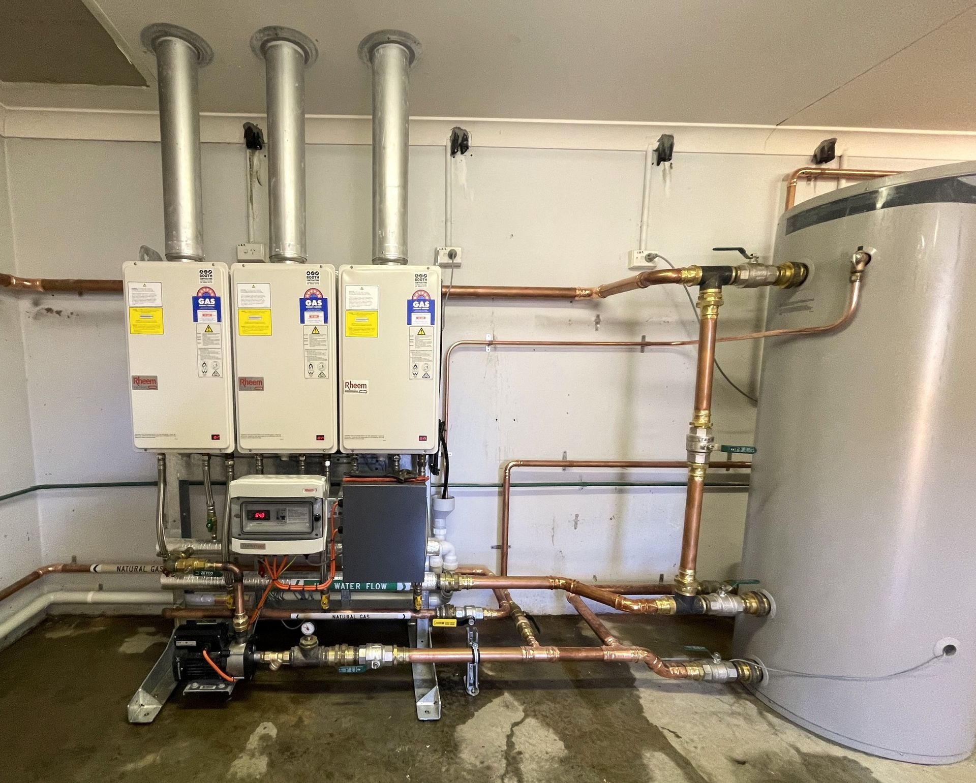 Mechanical room with three wall-mounted boilers, silver piping, and a large cylindrical tank on the right. Mechanical room with three wall-mounted boilers, silver piping, and a large cylindrical tank on the right.