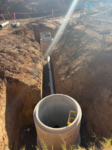 Excavated trench with a large concrete manhole ring and pipe sections in a construction site under sunlight