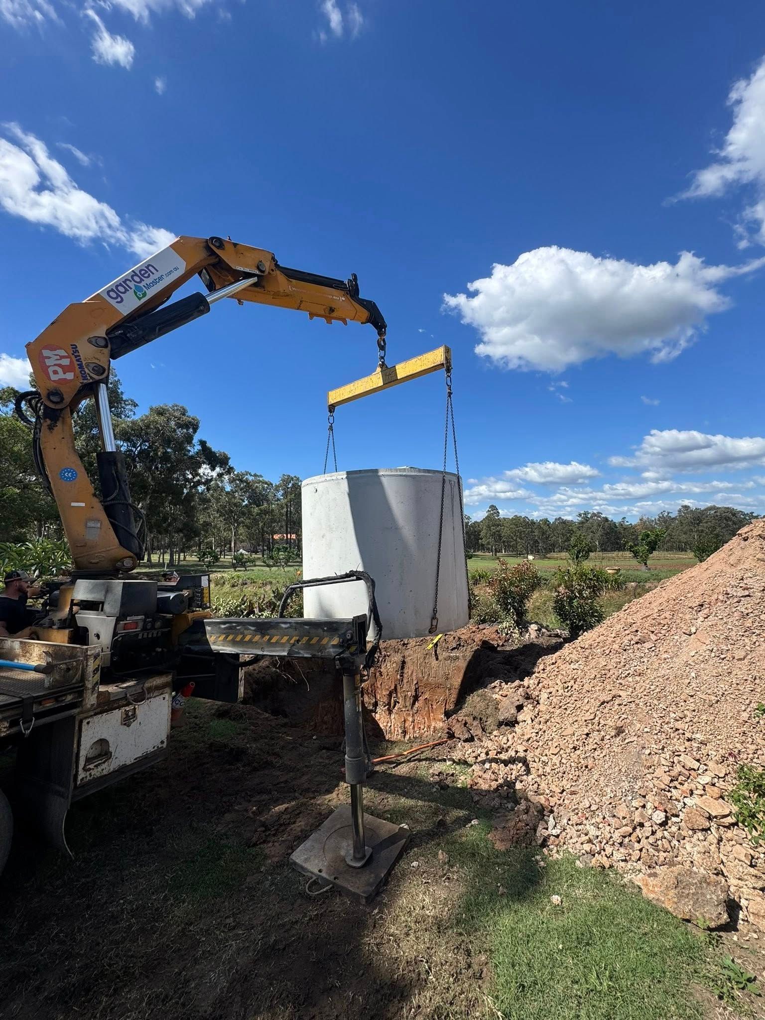 Excavator lifting a white concrete tank beside a large gravel pile under a blue sky Excavator lifting a white concrete tank beside a large gravel pile under a blue sky