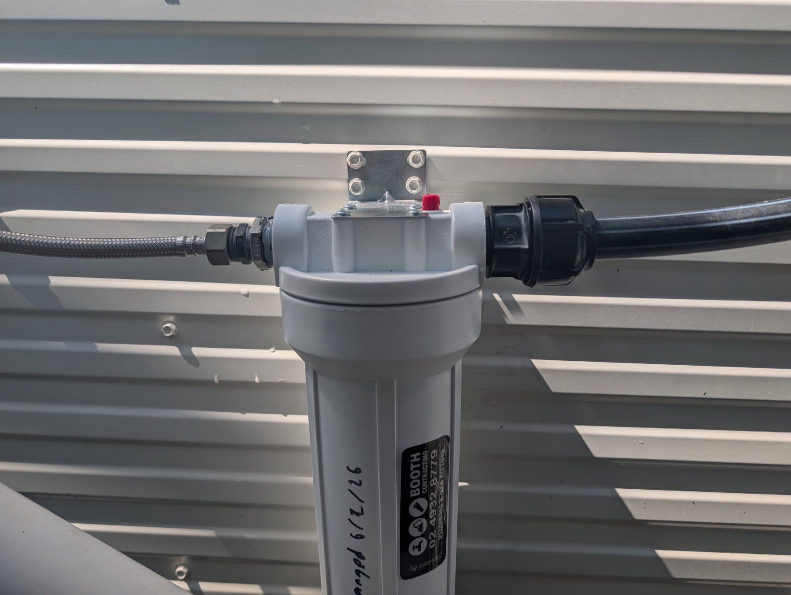 Gray inline water filter installed on a black pipe against a corrugated metal wall.