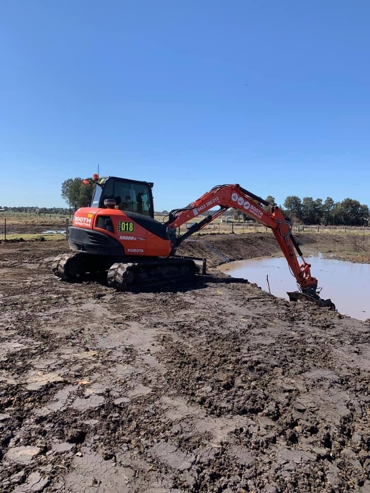 Orange excavator digging mud beside a small pond under a clear blue sky