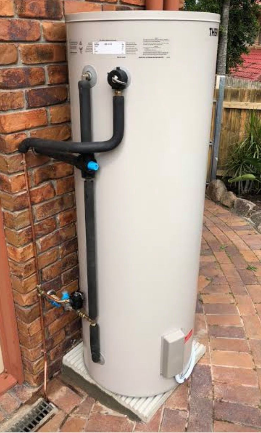 White cylindrical water heater beside a brick wall, with black pipes and a blue valve outdoors. White cylindrical water heater beside a brick wall, with black pipes and a blue valve outdoors.