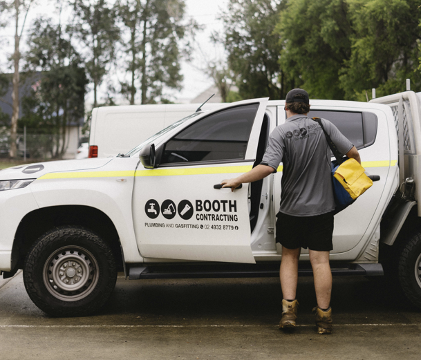 Man standing by a white Booth Construction pickup truck, loading a yellow bag in a rainy lot.