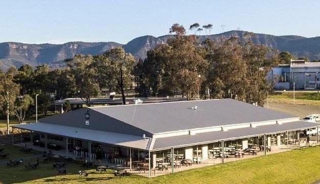 Single-story building with a gray roof in a grassy field, with trees and mountains in the background.