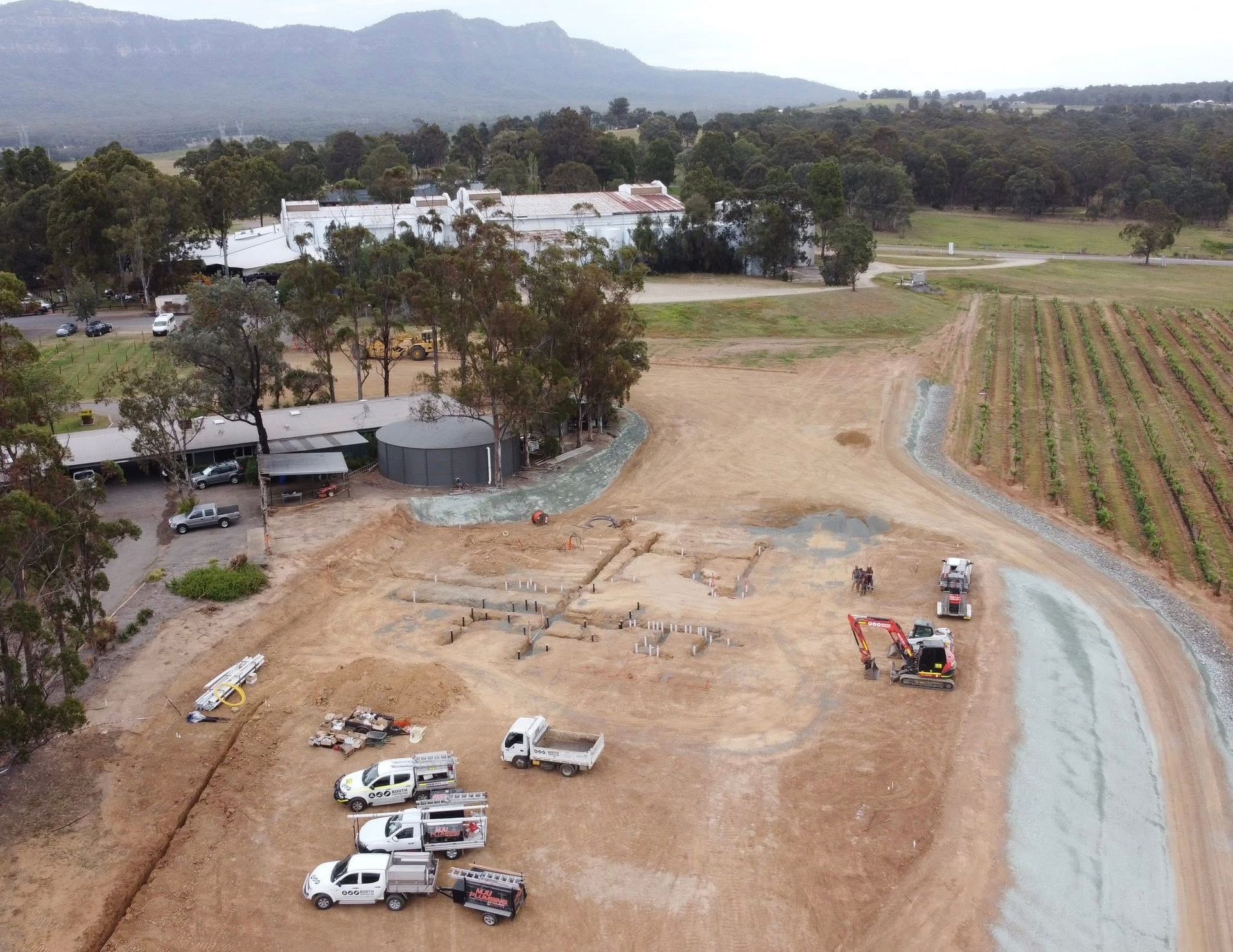 Aerial view of a rural construction site with trucks, dirt lots, a gravel road, and farm fields nearby