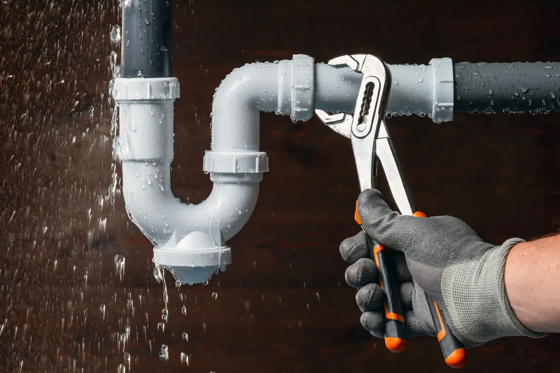 Gloved hand tightening a wrench on a leaking white pipe under a sink, with water dripping Gloved hand tightening a wrench on a leaking white pipe under a sink, with water dripping