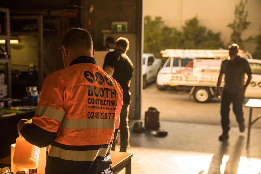 Worker in orange safety vest stands in a garage with another person and a service truck outside.