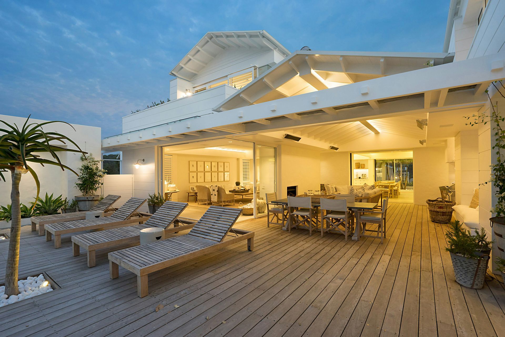 Sunlit seaside deck with lounge chairs, dining tables, and a modern white beach house at dusk