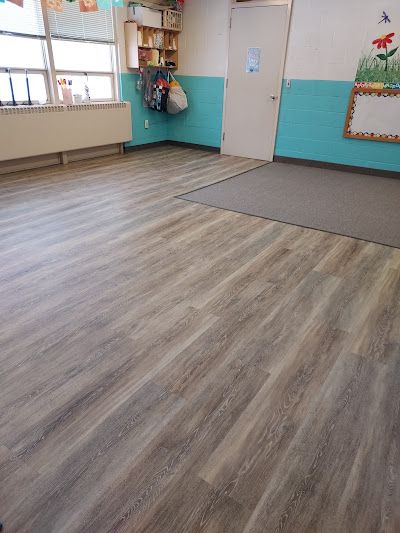 An empty classroom with wood-patterned flooring, a gray area rug, light blue walls, a white door, and a wall-mounted shelf.