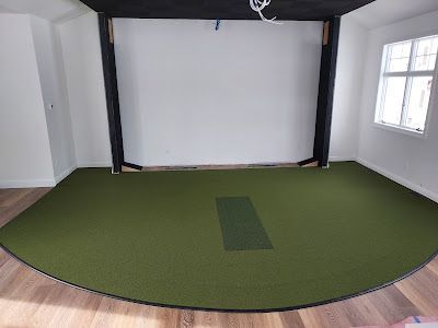 An indoor golf simulator room with a green turf floor, a central hitting mat, and a white screen framed by black beams.