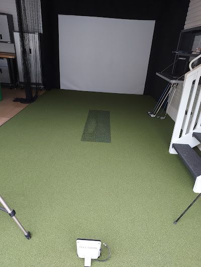 An indoor golf simulator setup with a white projection screen, green artificial turf, a hitting mat, and floor sensors.