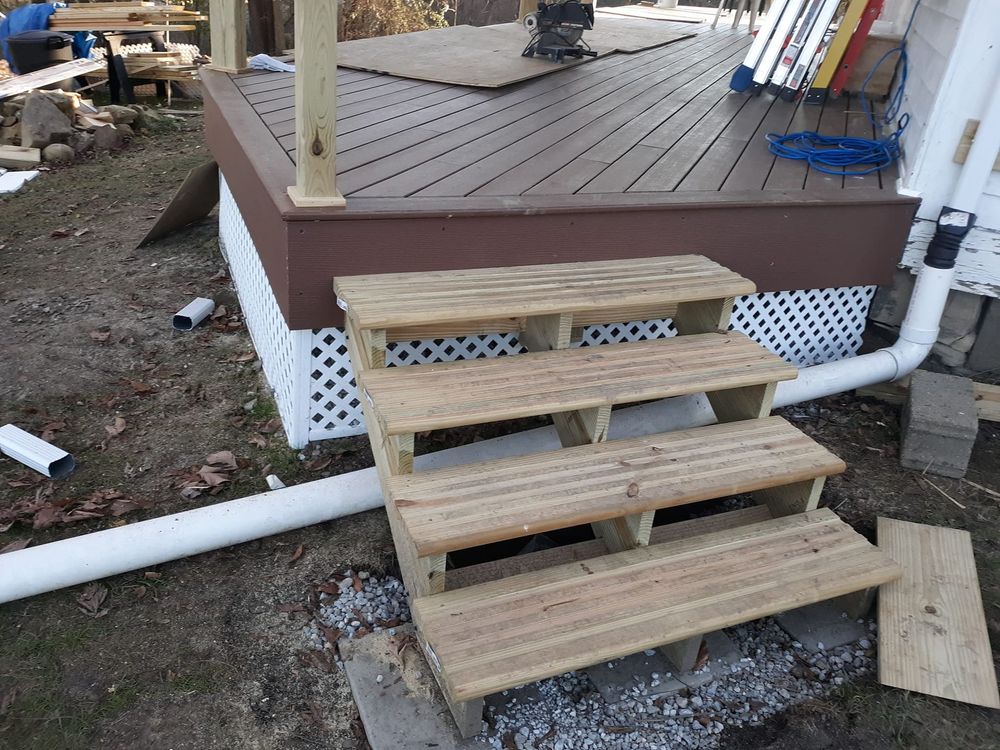A set of unfinished wooden stairs leading to a brown deck with white lattice skirting and a white pipe on the ground.