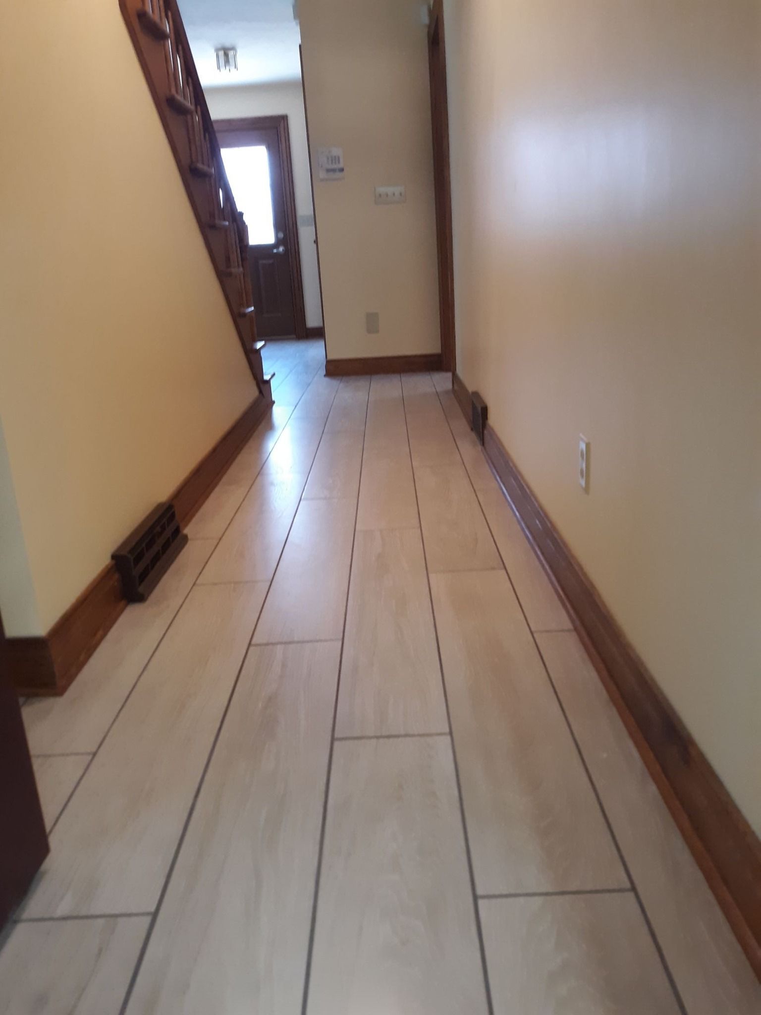 A hallway with light-colored, wood-look plank flooring, beige walls, and dark wood trim near a staircase.