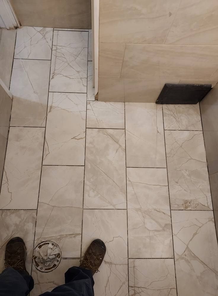 A first-person view looking down at a bathroom floor with newly installed beige marble-patterned tile around a toilet flange.