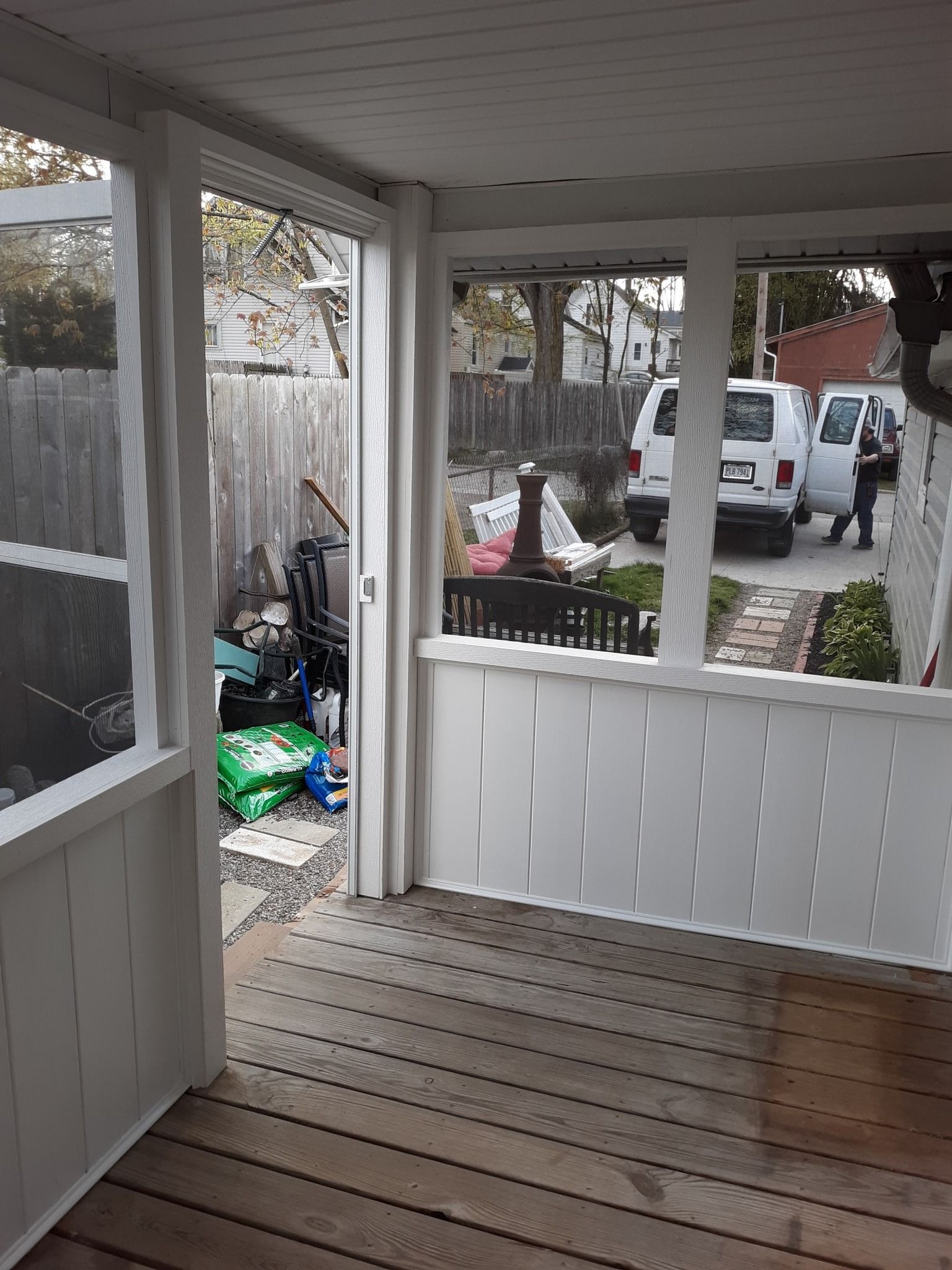 A screened-in porch with white wooden railings and a wooden floor, opening to a yard with a parked white van.