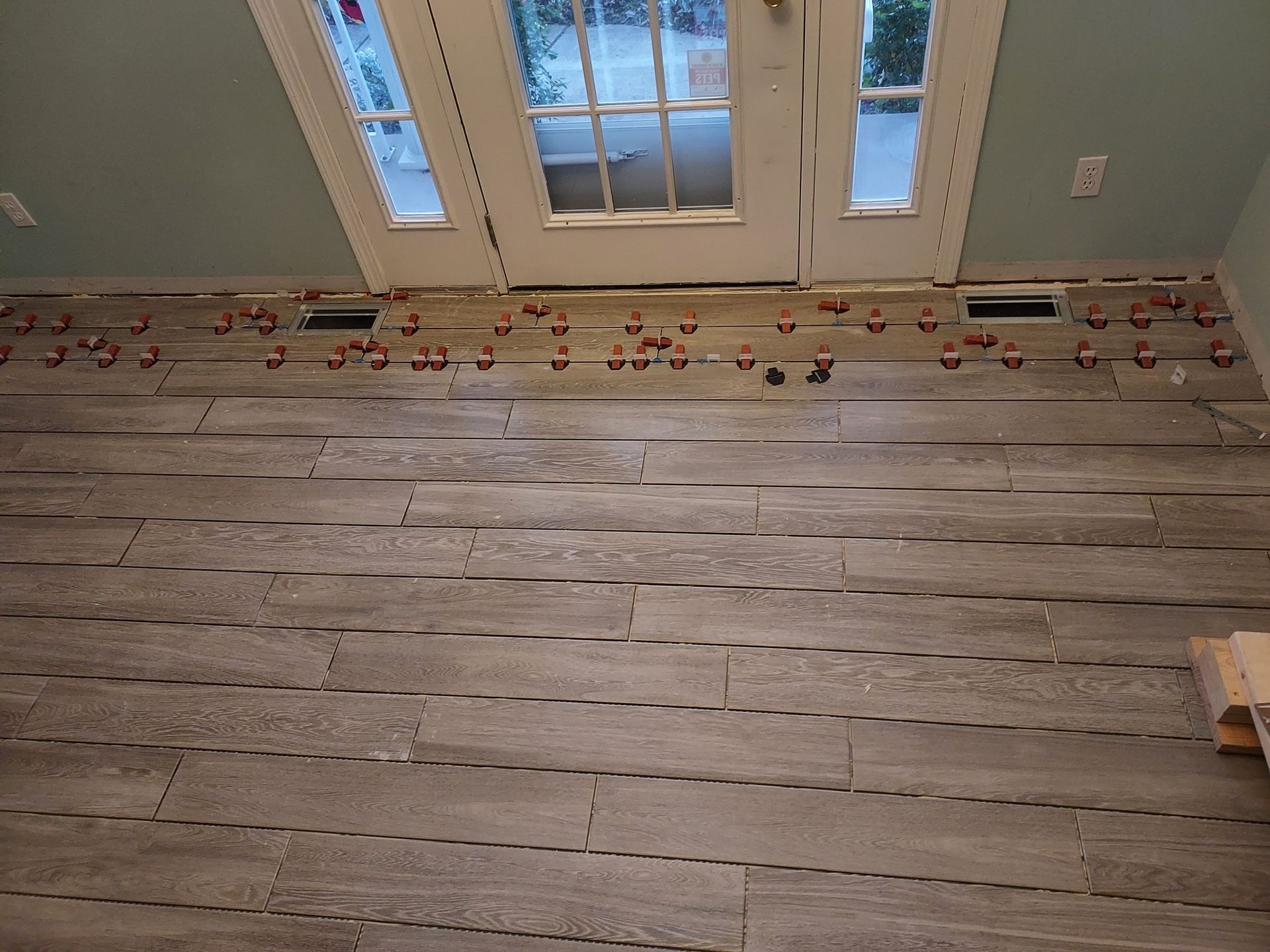 Newly installed gray wood-look vinyl floor planks with plastic leveling spacers inserted in the gaps, near a glass door.