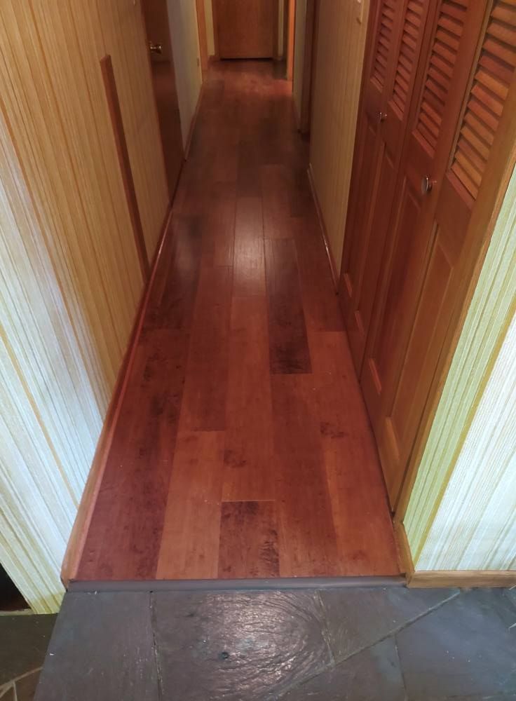 A transition from gray stone tile flooring to a long, narrow hallway with reddish-brown wood planks and light wood walls.