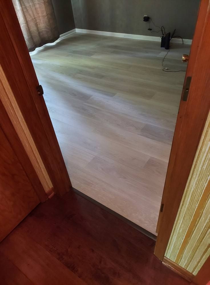A doorway frames a view into a room with light, neutral-toned flooring, contrasting with the dark wood floor in the hall.