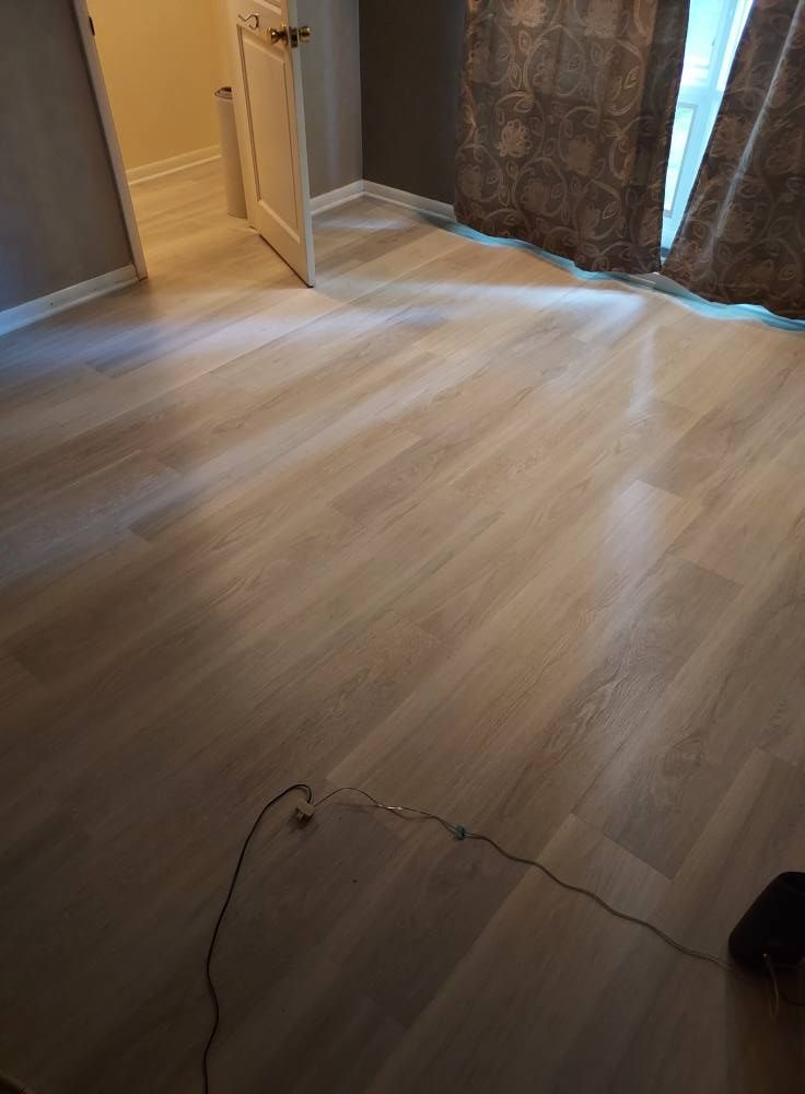 Light-colored wood flooring in an empty room, with a door open to the left and brown curtains hanging near a window.