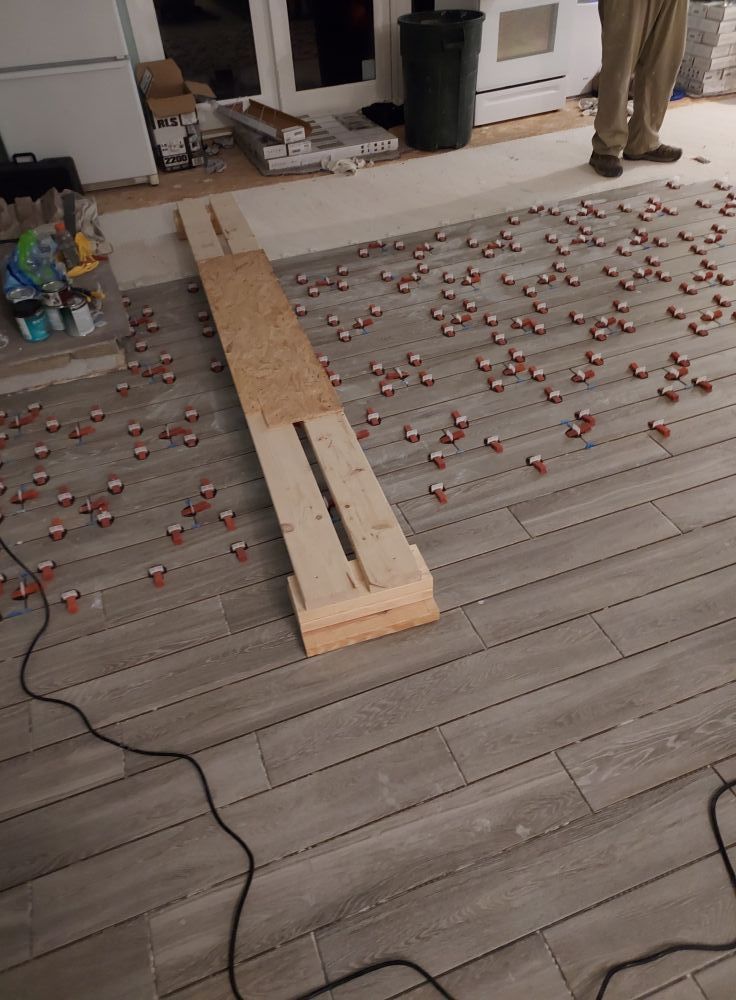 A floor installation project with gray wood-look tiles, red leveling spacers, and a wooden board for kneeling.