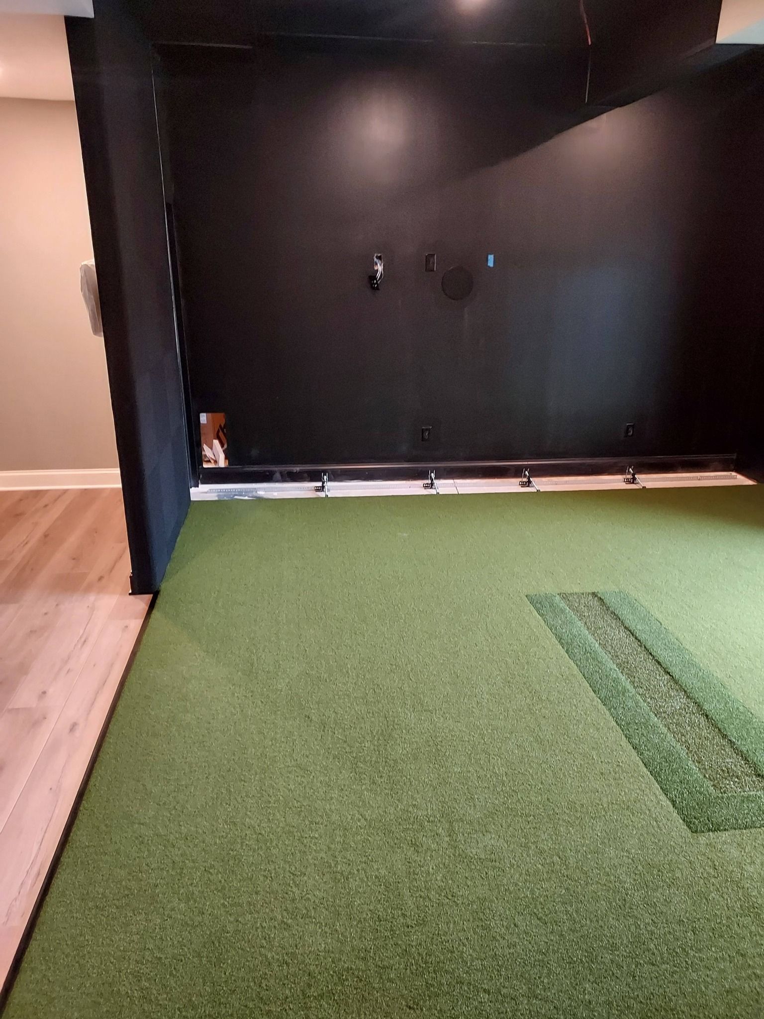 An indoor golf simulator room with green artificial turf, a hitting strip, and a dark black wall for a projector screen.