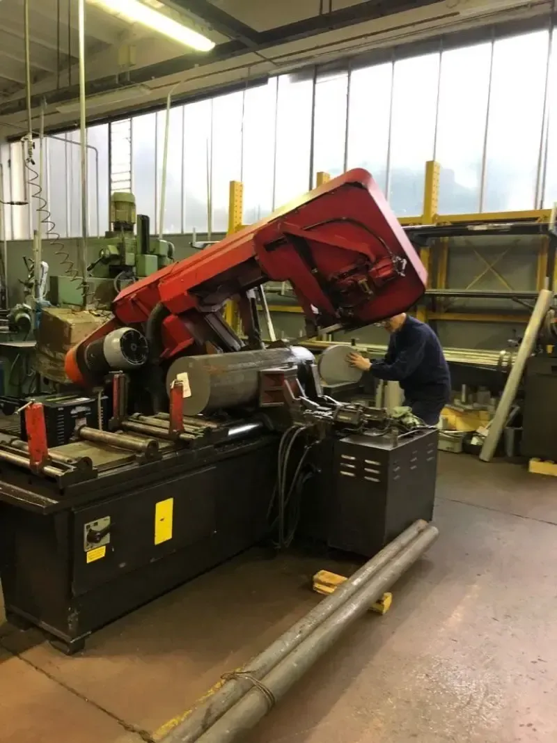 Una persona utilizza una sega a nastro rossa e nera in un'officina, con barre di metallo e altri macchinari nelle vicinanze.