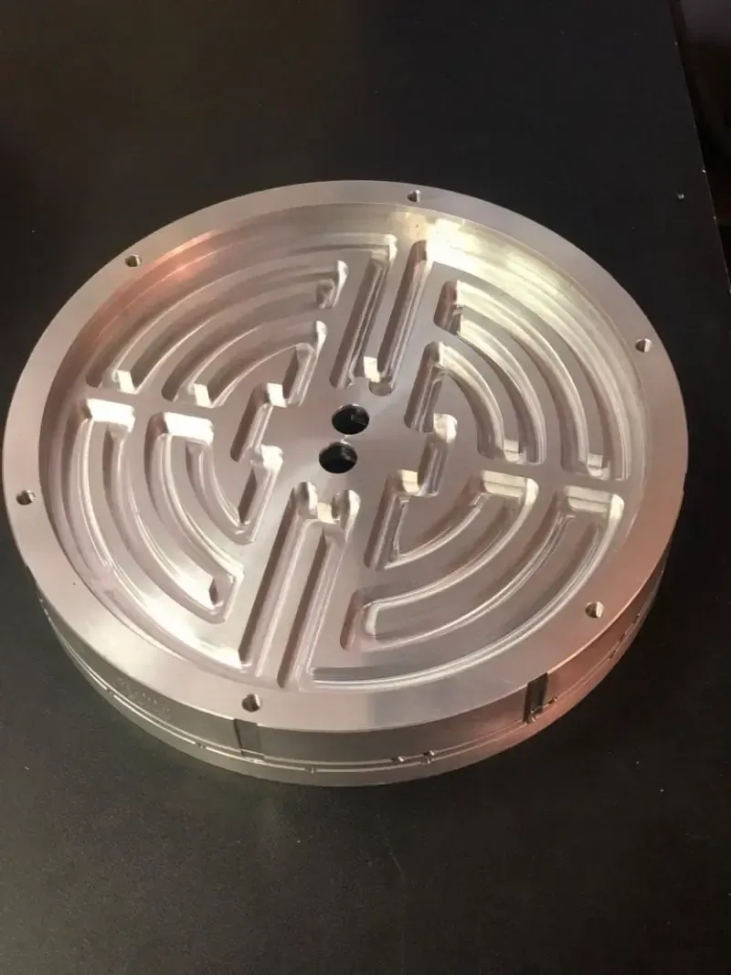 Componente metallico circolare con un intricato design a canale, con due piccoli fori al centro.