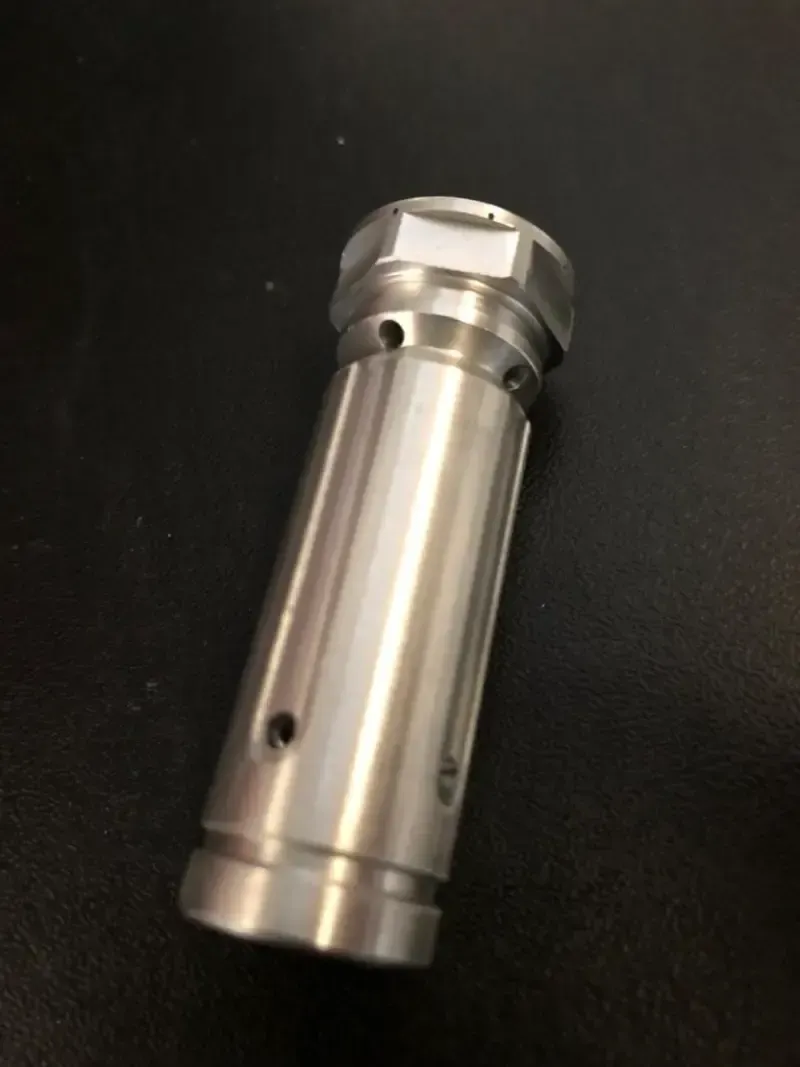 Pezzo industriale cilindrico in metallo lucido con parte superiore esagonale e diversi fori.