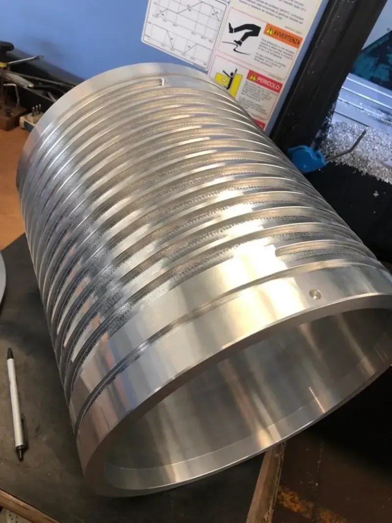 Pezzo metallico cilindrico con scanalature circolari. Si trova su una scrivania accanto a una penna e a una cianografia.