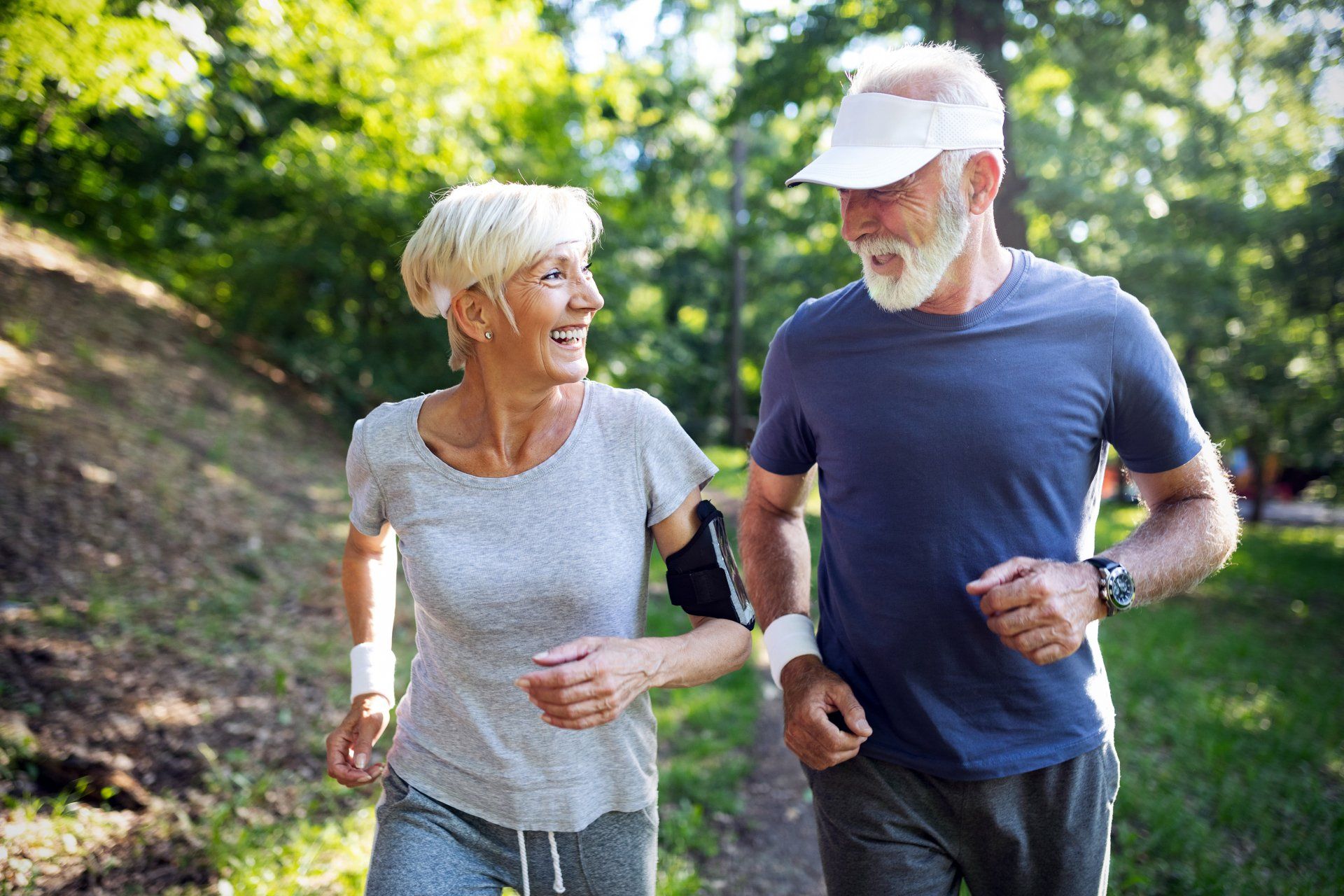 An older couple is jogging in the sunshine and smiling at one another.