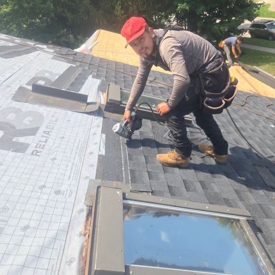 A roofer in a red cap using a nail gun to install asphalt shingles on a residential roof near a skylight.