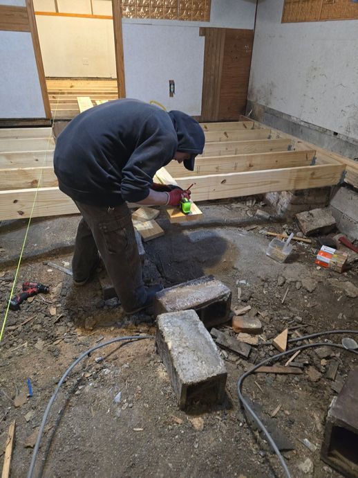 A person in a dark hooded sweatshirt marks a wooden floor joist with a red pencil while working in a room under renovation.