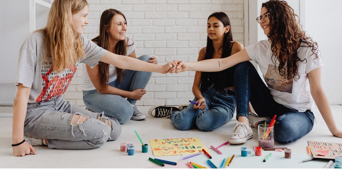Four people sit in a circle on the floor, holding hands, with art supplies scattered around them.